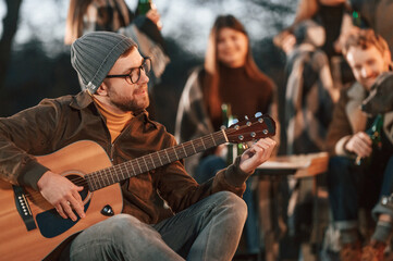 Obraz premium Man in hat is sitting and playing guitar. Group of people is spending time together on the backyard at evening time