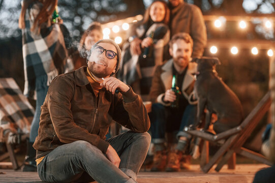 Man Is Smoking. Group Of People Is Spending Time Together On The Backyard At Evening Time