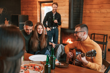 Man in glasses is playing guitar. Group of friends is having good weekend indoors in the wooden building together