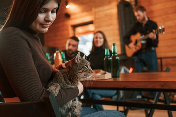 Woman with cat on her legs. Group of friends is having good weekend indoors in the wooden building together