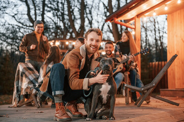 Happy man is sitting with his dog. Group of people is spending time together on the backyard at evening time