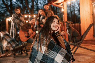 Young woman with a blanket is sitting. Group of people is spending time together on the backyard at evening time