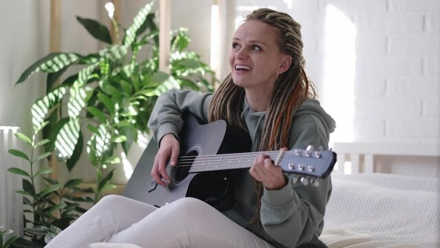 woman with dreadlocks on head singing song and playing acoustic guitar in sunny cottage bedroom, simple living and hobby relaxing