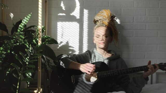 woman with dreadlocks on head singing song and playing acoustic guitar in sunny cottage bedroom, simple living and hobby relaxing