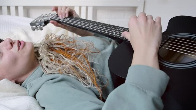 woman with dreadlocks on head singing song and playing acoustic guitar lying on bed in sunny cottage bedroom, simple living and hobby relaxing