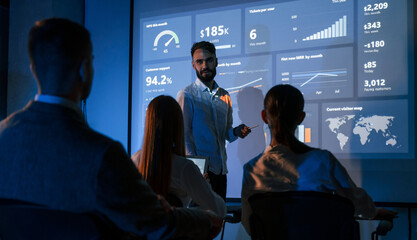 Male leader talking to employees, showing the plan on the projector in office of stock exchange...