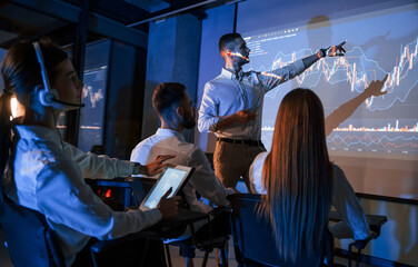 Company growth plan. Male leader talking to employees, showing information on the projector in...