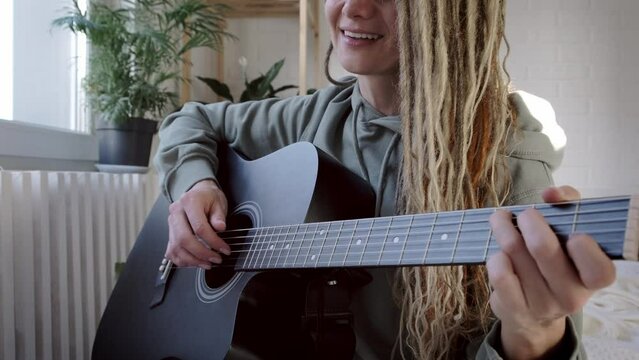 woman with dreadlocks on head singing song and playing acoustic guitar in sunny cottage bedroom, simple living and hobby relaxing
