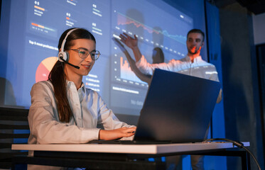 Stock broker is sitting and working by laptop. Man is standing near her, showing data on projector