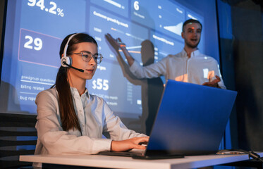 Stock broker is sitting and working by laptop. Man is standing near her, showing data on projector