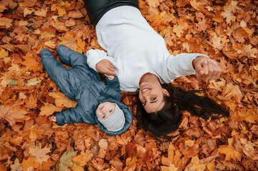 Woman with her little son is laying down on the ground that is covered with autumn fallen leaves
