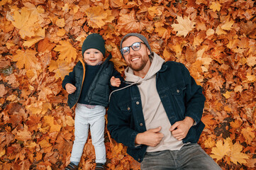 Laying down on the ground that covered with fallen leaves. Father and young son is together outdoors at daytime