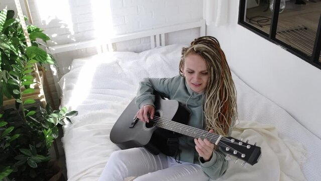 woman with dreadlocks on head singing song and playing acoustic guitar in sunny cottage bedroom, simple living and hobby relaxing