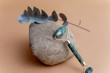 Obraz premium Green jade roller for facial massage on a brown stone with a dry branch of eucalyptus on a beige background. Natural anti-aging massage products for lymphatic drainage and wrinkle smoothing.Selective