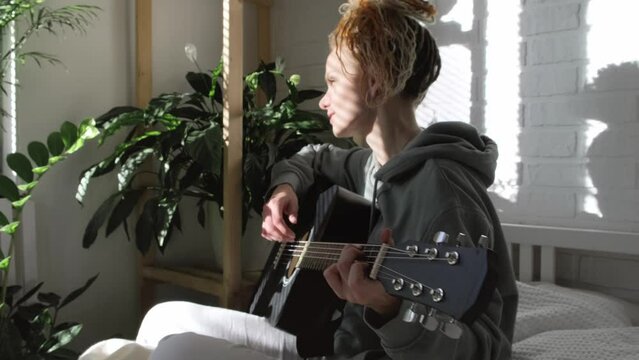 woman with dreadlocks on head singing song and playing acoustic guitar in sunny cottage bedroom, simple living and hobby relaxing
