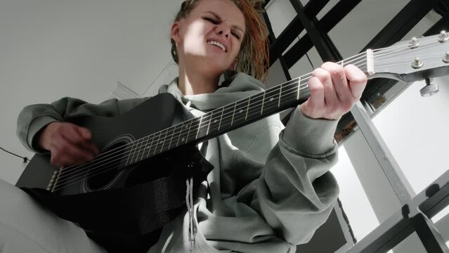 woman with dreadlocks on head singing song and playing acoustic guitar in sunny cottage bedroom, simple living and hobby relaxing