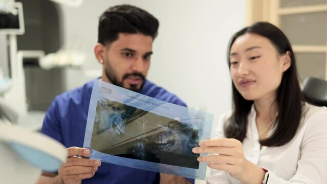 Focus on x-ray image. Medicine, dentistry and oral care concept. Male confident dentist showing x ray picture to attractive young patient, asian woman at modern light dental clinic.