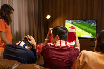 Group of Asian people friends sit on sofa watching and cheering football or soccer games competition on TV together at home.Happy man and woman sport fans celebrating sport team victory sports match
