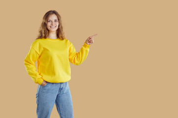 Friendly smiling young blondy woman with wavy hair wearing yellow sweatshirt and jeans pointing index finger on copy space on right on beige background looking at camera keeping one hand in pocket.