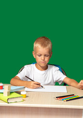 Boy looks attentively at piece of paper with pencil in his hand. There are pencils, book, cubes, paints on table. Free time, learning to draw, hobbies. Selective focus.