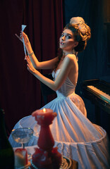 Fototapeta premium Portrait of beautiful young woman in image of medieval person, musician in tender white dress sitting at piano with new music composition. Comparison of eras, beauty, history, art