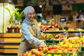 Female seller in hijab browsing and checking apples in supermarket, woman in apron smiling at work in store in fruit and vegetable department.