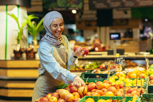 Female seller in hijab browsing and checking apples in supermarket, woman in apron smiling at work in store in fruit and vegetable department. - Powered by Adobe