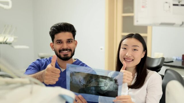 Smiling happy asian woman visiting dentist, sitting in dental chair at modern light hospital clinic. Young bearded man dentist holding x ray image scan showing thumb up.