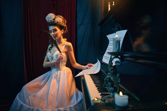 Portrait of beautiful young woman in image of medieval person in tender white dress sitting at piano with quill and notes. Making lovely melody. Comparison of eras, beauty, history, art