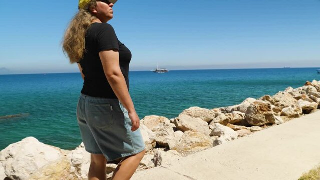 Woman in shorts, jersey and baseball cap walking along seafront. The camera is moving in parallel. The woman has an action camera in her hand. In background is a beautiful sea, mountains and boats