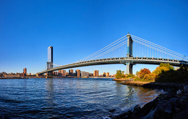 Panoramic view from Brooklyn of iconic Manhattan Bridge over blue waters leading to New York City with blue sky