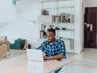 African American man in glasses sitting at a table in a modern living room, using a laptop for business video chat, conversation with friends and entertainment