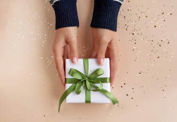 Female hands in sweater with pink manicure holding present in white paper with satin ribbon on festive beige background. Xmas composition. Flat lay. Happy holidays, New Year celebration concept. 