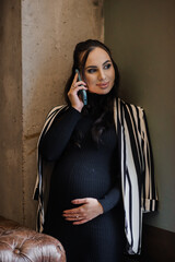 Fototapeta premium Beautiful young pregnant woman in black dress and black and white jacket talking on smartphone