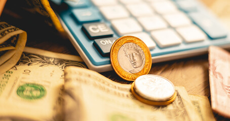 Uma moeda do Real Brasileiro de 1 Real sobre um móvel de madeira com cédulas de dólares americanos e um calculadora na composição da imagem. Conceito de câmbio, negócios internacionais e inflação.