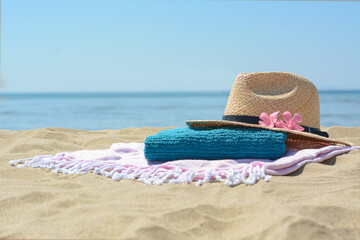 Fototapeta na wymiar Blanket with towel, stylish straw hat and flowers on sand near sea, space for text. Beach accessories