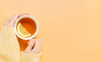 Female hands in yellow sweater hold cup of tea with lemon, copy space, orange background