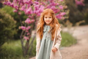 Smiling little kid girl 4-5 year old wear stylish beige jacket and dress with ginger long hair walk in park over flowers outdoor. Springtime. Childhood.