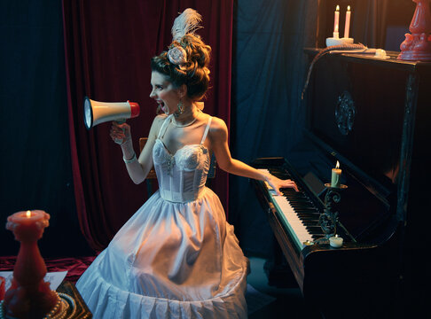 News. Portrait of young beautiful girl in image of medieval person in elegant white dress sitting at the piano and shouting at megaphone. Comparison of eras, beauty, history, art