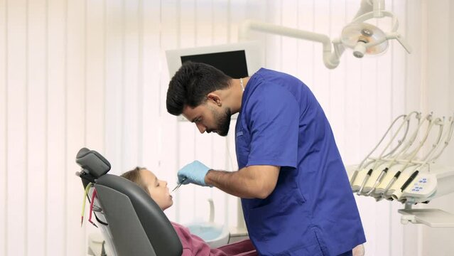 Side view of smiling male professional doctor in blue uniform and gloves, making examination or curing caries and toothache for his little patient, cute kid girl, sitting in dental chair.