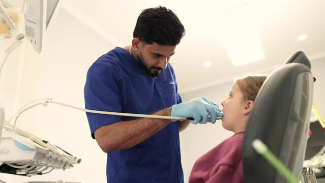 Shot of young confident bearded dentist in blue uniform, making dental check up and caries treatment with drill for her cute little patient, little girl, sitting in dental chair at clinic.