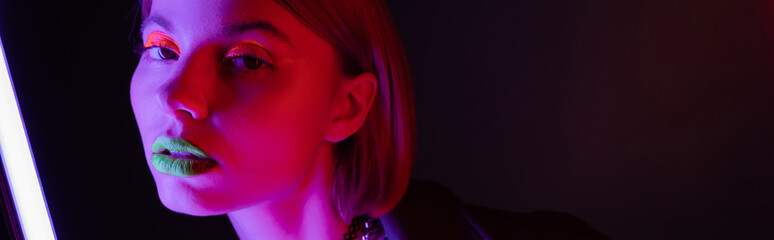 Fototapeta premium portrait of young woman with neon makeup posing in purple light of neon lamp on black background, banner.