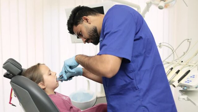 Cute pretty little girl during the dental procedure at the modern children's dental clinic. Children's attractive male bearded dentist curing teeth of a girl, using dental drill and mirror.