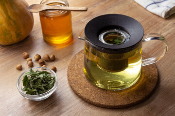 Glass teapot with green mint tea on wooden table with honey, pumpkin and hazelnut.