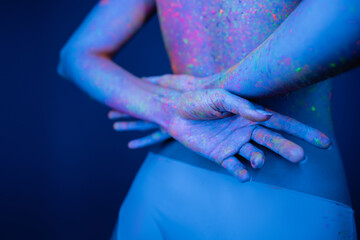 cropped view of young woman in bright neon paint holding hands behind back isolated on dark blue.