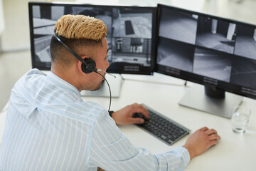 High angle view at Asian man wearing headset looking at multiple surveillance feeds in monitoring and security center