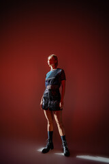 full length view of woman in leather boots and mini skirt standing in sunlight on burgundy background.