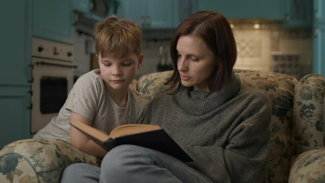 Mother and kid reading paper book. 30s woman enjoys reading literature with her son sitting on cozy sofa at home.