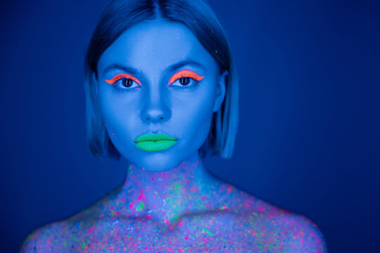portrait of woman in vibrant neon makeup and fluorescent body paint looking at camera isolated on dark blue.
