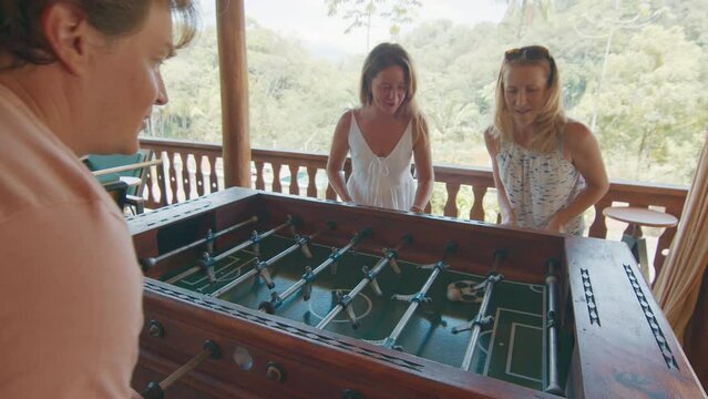 Two women and man play table football on the balcony. Girls playing foosball in the garden and celebrate goal